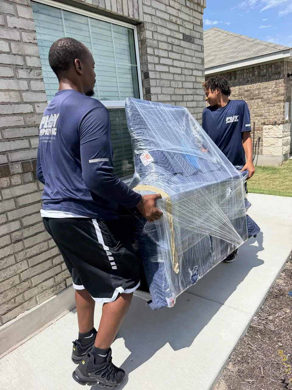 Two Playhouse Moving crew members carrying a padded and wrapped chair beside a brick home